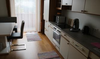 A modern kitchen with a light wood paneling and a large window. The room is equipped with a dining table and chairs as well as various kitchen appliances.