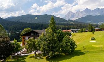 A picturesque landscape with a hotel surrounded by trees and meadows. In the background, majestic mountains rise under a blue sky.