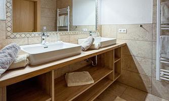 A modern bathroom with two sinks and wooden details. The walls are covered with tiles and a large mirror provides brightness.
