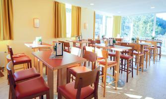 A bright and inviting restaurant with wooden chairs and tables.  
Large windows offer a view of the surroundings and create a pleasant atmosphere.