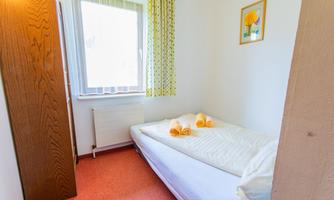 A simple room with a single bed and fresh towels. A window with bright curtains allows for daylight.