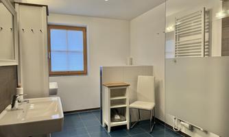 A modern bathroom with blue tiles, a sink, and a glass shower partition. There is a bright wall arrangement and a chair next to a small shelf.