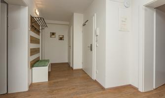 A bright hallway with wooden flooring and white walls. On the left side, there is a bench and a coat rack panel.