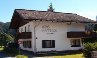 A charming guesthouse named "Haus Kampenblick" in nature. It has a traditional Alpine style with a balcony and is surrounded by green trees.