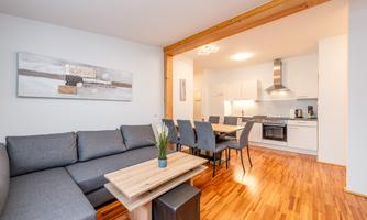 A modern living room with a gray sofa and a wooden table. In the background, there is an open kitchen with white cabinets and a dining table.