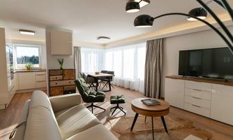 A modern living room with a light hardwood floor and plenty of natural light. There is a cozy couch, a round table, and a television.