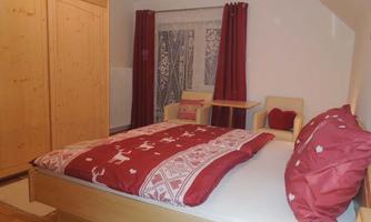 A cozy bedroom with a wooden bed and red pillows. The walls are light, and there is a wardrobe as well as a small sitting area.