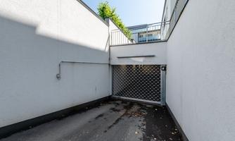 Access to an underground garage with a grille gate. The walls are white and the surroundings are sunny with some green foliage visible.