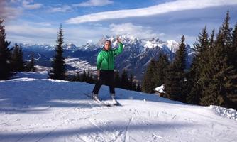 A skier stands on a snow-covered slope and waves. In the background, impressive mountains and a clear blue sky can be seen.