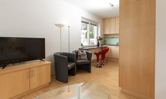 A modern living room with wooden flooring and a large window front. The room is furnished with a television, two black bar stools, and one red bar stool.