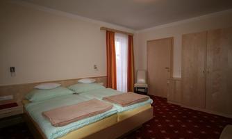 A simple bedroom with two beds, a table, and a chair. The room has a warm carpet and large windows with curtains.