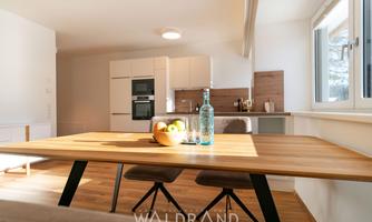 A modern kitchen with a wooden table and two chairs. On the table, there is a fruit bowl and a glass bottle of water.