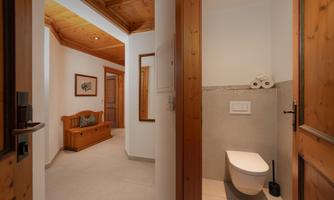 A modern hallway with a toilet in the foreground and bright walls. On the left, there is a wooden bench and a wall mirror.