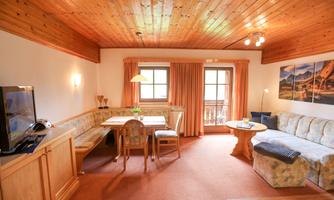 A cozy living space with wooden walls and a large window. The room features a sofa area, a dining table, and a television.