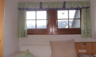 A room with a window that offers a view of the mountains. The curtains are green with a cheerful pattern, and a cozy sofa is placed by the window.