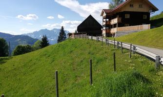 A green meadow with a house in the background and mountains in the distance. The sky is blue with some clouds.