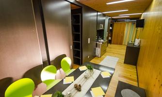 A modern room with wooden paneling and bright light. A table with colorful chairs and a small kitchenette is visible.