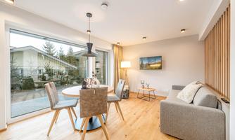 A modern living room with a round dining table, chairs, and a cozy sofa. Large windows offer a view of the outdoor area.