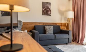 A cozy living room with a gray sofa and a wooden table. The walls are light, and there is a floor lamp as well as curtains.