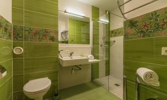A modern bathroom with green tiles and a shower cabin. There is a sink, a toilet, and fresh towels.