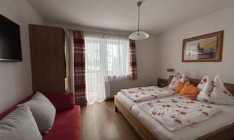 A cozy bedroom with two beds and colorful pillows. Large windows let in plenty of light, and there is a small living area with a couch.
