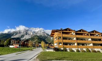 A picturesque mountain landscape with majestic peaks and a modern wooden building in the foreground. The clear sky and lush meadows give the scene an inviting atmosphere.