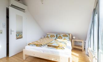 A cozy bedroom with a double bed and wooden flooring. There is a large window area that lets in plenty of daylight.
