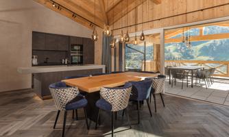 A modern kitchen with wood decor and a large dining table. Large windows offer a view of the mountains and plenty of natural light.