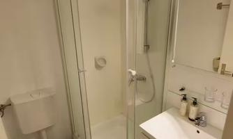 A modern bathroom with a transparent shower cabin and a sink. There are personal care products on the sink and a toilet is available.