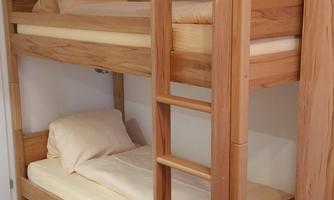 A simple wooden bunk bed in a modern room. Below it is a single bed, and the floor is clad in wood.