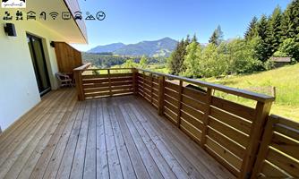 A spacious balcony with wooden flooring and a view of green hills and forests. The sky is clear and sunny, ideal for relaxing moments outdoors.