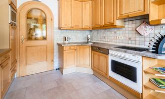 A bright kitchen with wooden cabinets and modern appliances. The floor is tiled, and there is a glass door that leads to additional living space.