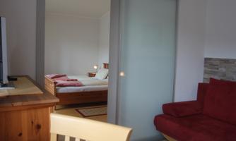 A cozy apartment with a red sofa and a table. In the background, a bedroom with a double bed is visible.