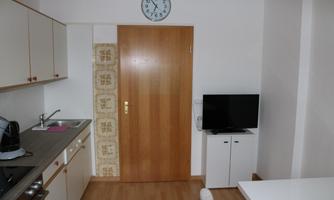 A small, bright kitchen with a table, a television, and a wooden shelf. On the wall hangs a clock and a wooden door leads to another room.