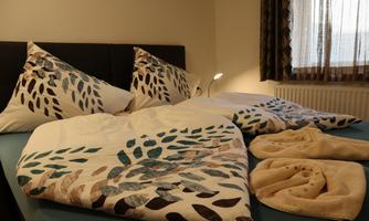 A cozy bedroom with a bed decorated with colorful cushions and a blanket. Next to the bed, there are two towels.