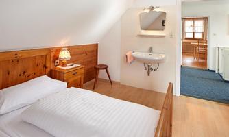 A cozy bedroom with a wooden bed and a small nightstand lamp. In the background, there is a small sink and access to another room.