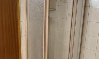 A shower with glass walls in a bright bathroom, separable by a shower partition. A white bench stands next to the shower.