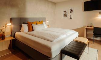 A modern bedroom with a large bed, cozy decor, and warm color tones. Pictures hang on the wall, and a table with a chair is also present.