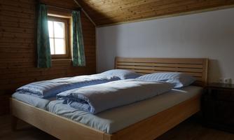 A cozy bedroom with a double bed and blue bedspreads. The walls are made of wood and there is a window with green curtains.