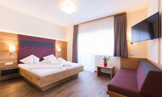 A modern hotel room with wooden decor, a double bed, and plenty of natural light. There is a cozy seating area and a TV on the wall.