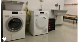 A laundry room with a washing machine and a dryer. In the background, there is a sink and a wooden bench.