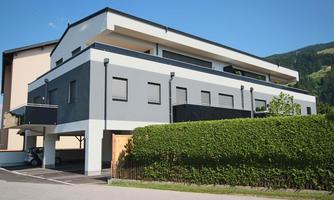 A modern apartment building with a gray facade and clean lines. In the foreground, a well-maintained hedge and a parking lot can be seen.