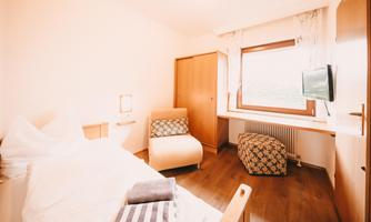 A cozy room with a single bed, a comfortable armchair, and a window that lets in plenty of light. There is also a small television and a rustic wooden dresser.