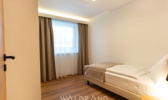 A simple, modern room with a single bed and wooden floor. Soft, natural lighting from a window with curtains.