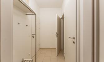 A bright hallway with white walls and a tiled floor. There are coat hooks on the wall and a door leads into another room.