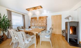 A cozy dining area with a long wooden table and chairs made of woven material. In the background, there is a decorative cabinet and a fireplace.