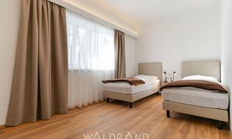 A modern room with two single beds and a large window. The walls are white and the floor is made of wood.