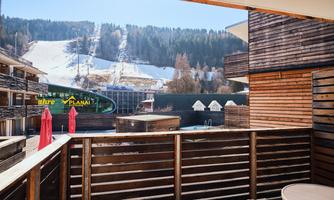 A view from a balcony overlooking a ski slope and a modern accommodation. In the background, snow-covered mountains and a blue sky can be seen.