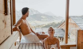 A relaxing moment in a sauna with a view of the mountains. Two people enjoy the tranquility in a wood-paneled setting.
