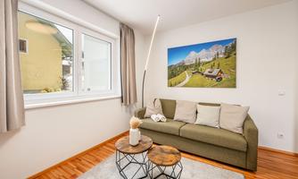 A cozy living room with a green sofa and two small tables. On the wall hangs a picture of the mountains and a alpine cabin landscape.